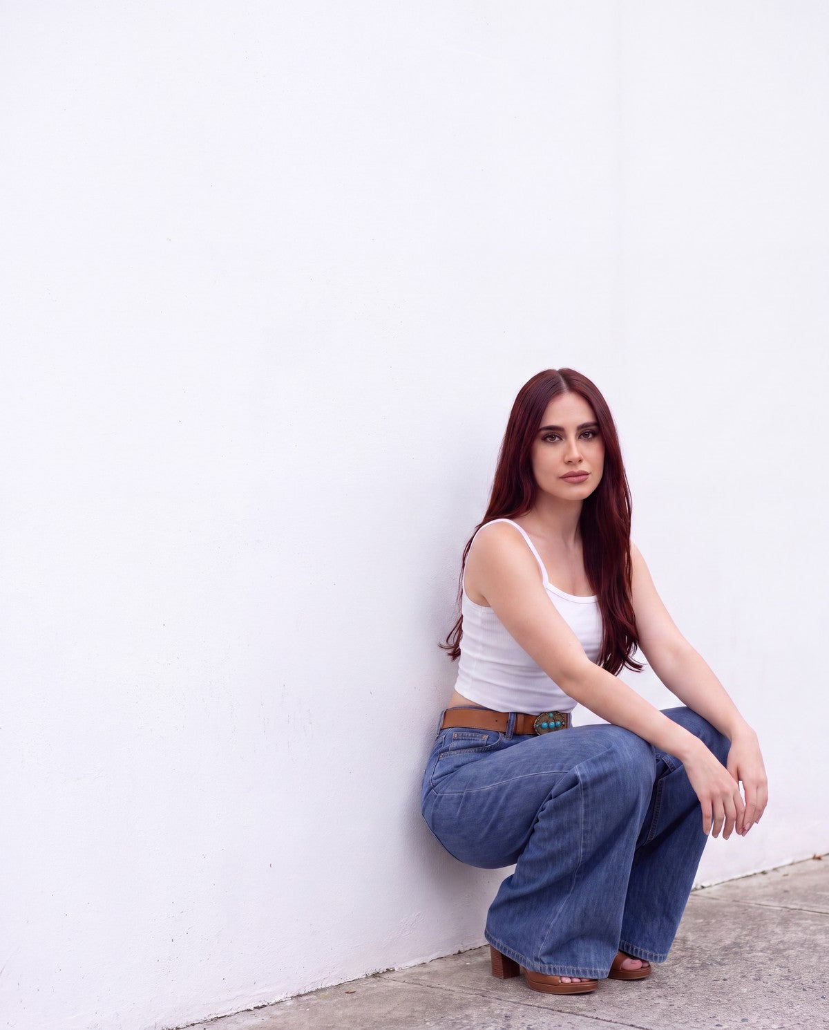 Jeans loose wide de mezclilla media con cinturón café, agachada junto a una pared blanca